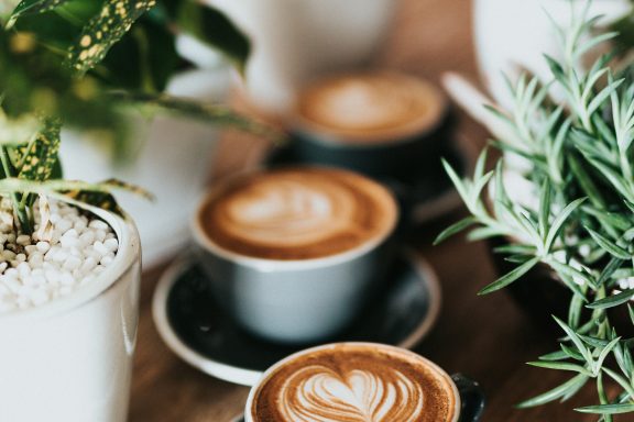 Drei Tassen Kaffee auf einem Holztisch, umgeben von Pflanzen.